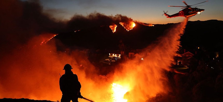 Fire personnel respond to homes destroyed while a helicopter drops water as the Palisades Fire grows in Pacific Palisades, California on January 7, 2025.