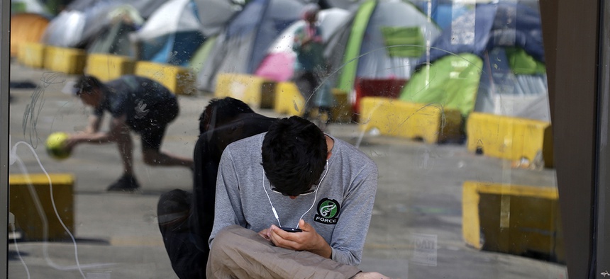 A migrant checks his cell phone as tents are reflected on a building window at the Athens port of Piraeus, Thursday, April 28, 2016.